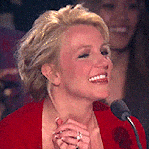 A smiling woman in a red top sits at a judges' table with a microphone, looking pleased.