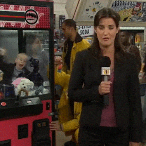 A TV reporter holding a microphone reports in front of an arcade claw machine.
