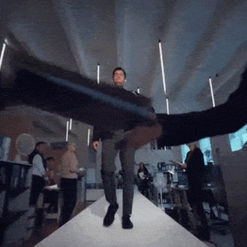 A man walks down an office aisle as coworkers keep piling papers and folders into his arms.