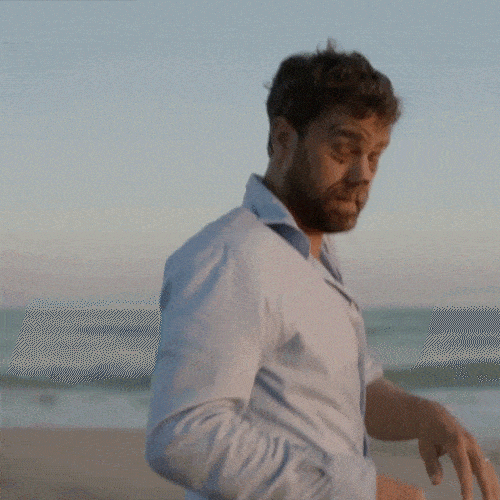 A man in a light blue shirt walks unsteadily on a beach at dusk. 