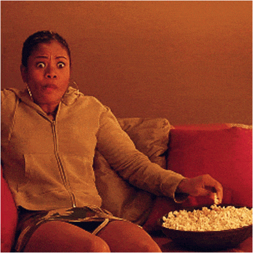 A woman on a red sofa with a bowl of popcorn covers her mouth in shock while watching something.