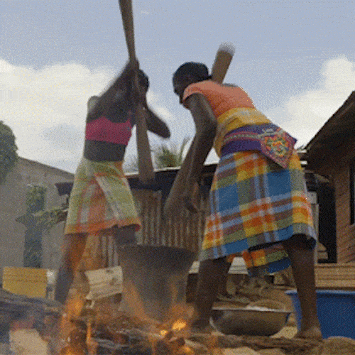 Two people pound grain with long wooden poles in a large mortar outdoors.