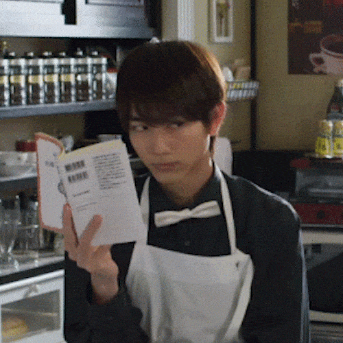 A young man in a waiter's apron reads a small booklet in a caf&eacute;.