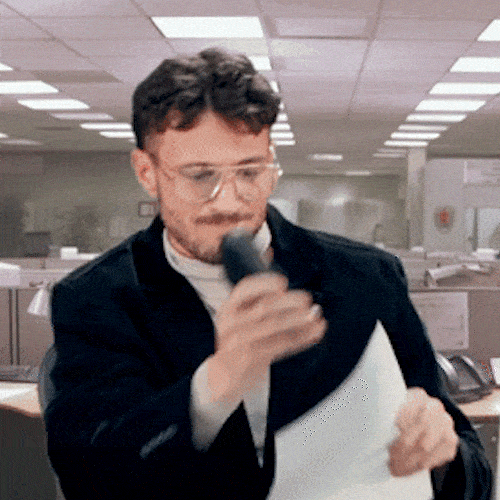 A man in an office holds up a sheet of paper and speaks into a microphone.