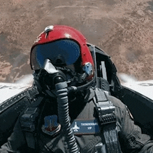 A fighter pilot in a helmet and oxygen mask sits in a cockpit before a jet flies across the sky.