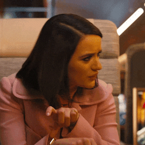 A woman in a pink coat taps her wrist as if pointing to a watch.