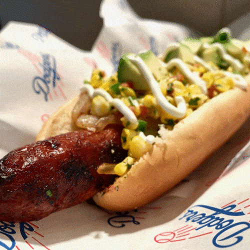 A grilled sausage and a hot dog topped with corn, avocado, and creamy sauce sit in a paper tray.