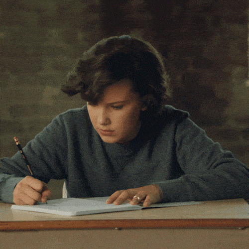 A girl writing at a desk looks up wide-eyed, as if about to say something.