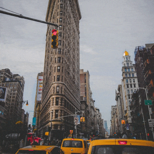 Yellow taxis drive past a tall triangular building on a busy city street.