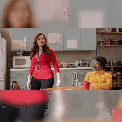 A woman in a red top strikes a confident pose in an office kitchen as a coworker watches.