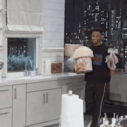 A smiling man holds a large box of white roses and a gift bag in a kitchen.