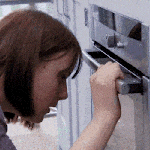 A woman crouches in front of an oven, watching closely through the glass door, then rests her head against it.