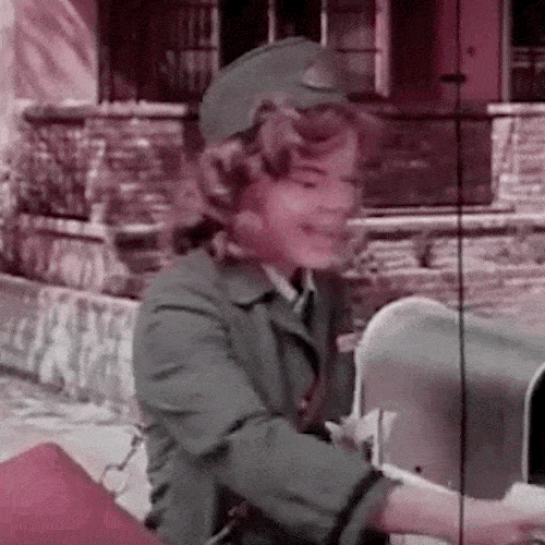 A smiling mail carrier in uniform with a shoulder bag delivers letters to a mailbox.