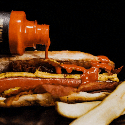 Hot sauce pours onto a stacked sandwich containing a hot dog, egg, and bacon, with fries beside it.