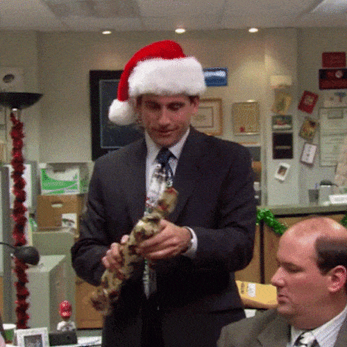 A man in a Santa hat and suit happily unwraps a gift in an office.