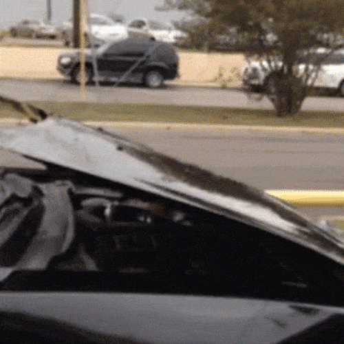 A popped car hood in the foreground with parked cars and a road visible behind it.
