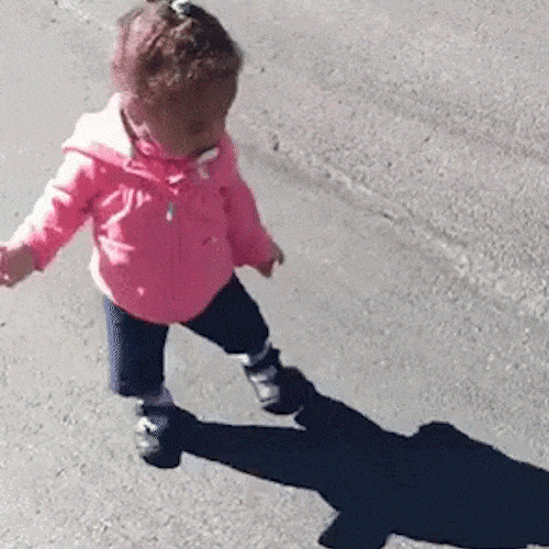 A toddler in a pink jacket walks along the pavement, followed by her shadow.