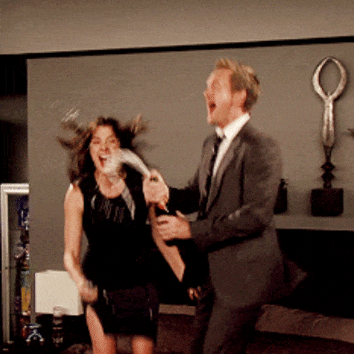 A man and woman in formal attire celebrate excitedly as he opens a bottle of champagne.