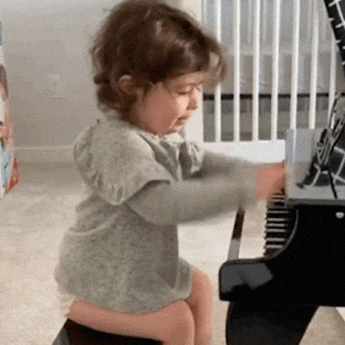 A toddler with curly hair plays a piano and smiles.