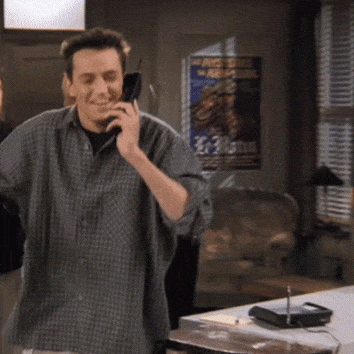 A man in a plaid shirt smiles while talking on a flip phone in a living room.
