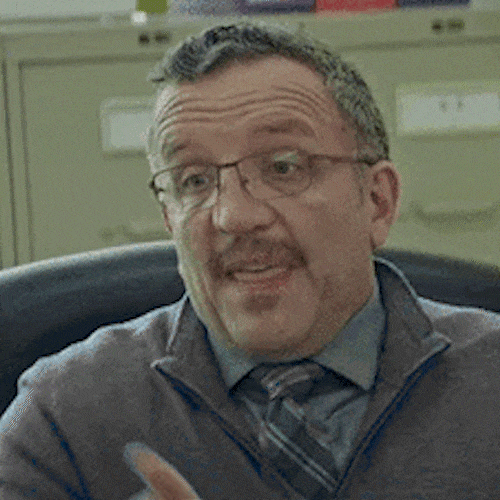 A man in glasses points off to the side while talking.