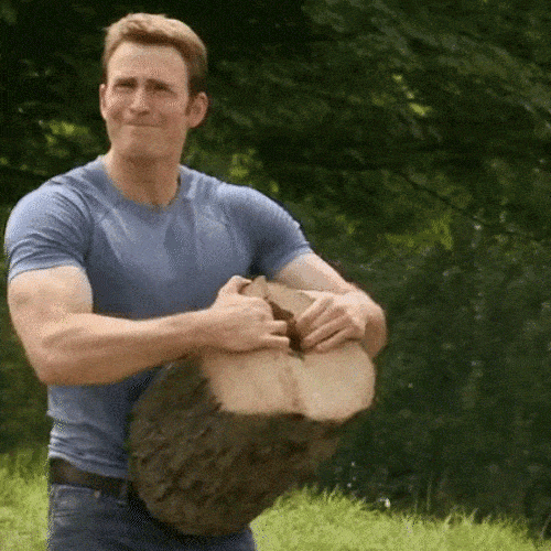A muscular man in a blue shirt breaks apart a large log outdoors.
