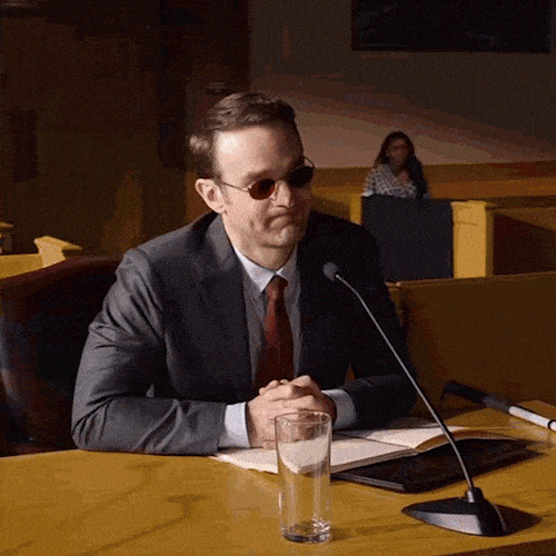 A lawyer in sunglasses and a suit sits at a hearing table with a microphone.