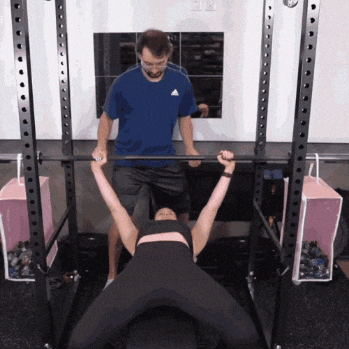 A man spots a woman lifting a barbell above her chest on a bench press.