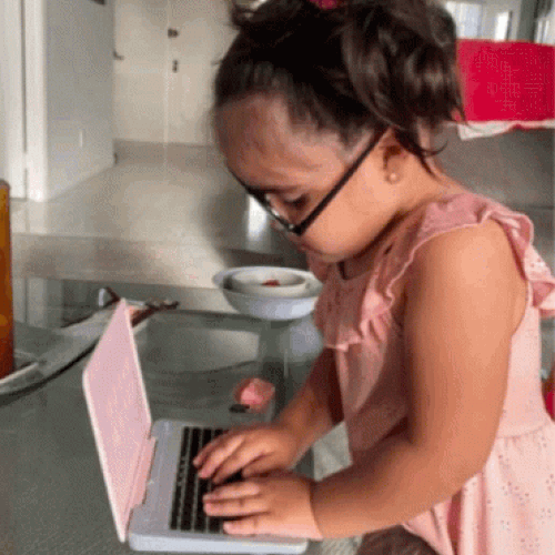A girl in glasses types quickly on a pink laptop.