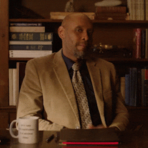 A man in a tweed jacket sits at a desk in a study and gives two thumbs-up.