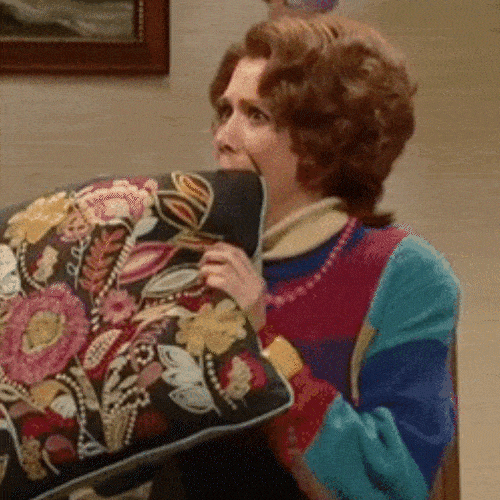 A woman with red hair hugs a large patterned cushion and bites it nervously.