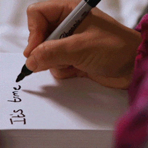 A hand writes with a black marker on paper, and a person in a tie-dye shirt writes at a desk.