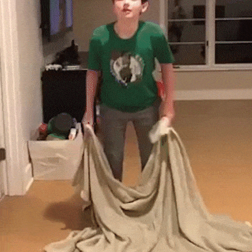 A boy holds up a blanket as if to make himself disappear, then falls to the floor.
