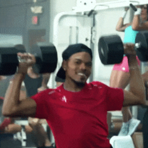 A man in a red shirt lifts dumbbells in each hand at the gym, with other people exercising in the background.