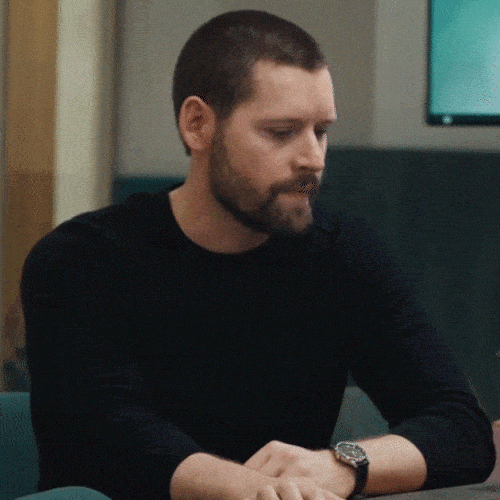 A man in a black sweater sits at a table looking down at his watch.