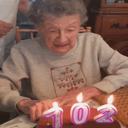 An elderly woman blows out candles shaped like the number 102 and laughs.