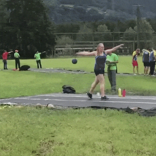 A hammer thrower spins on a sports field and lets go of the hammer, which flies low toward a man standing nearby.