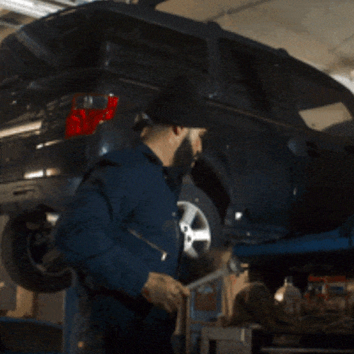 A mechanic in overalls stands beside a car raised on a lift in a garage.