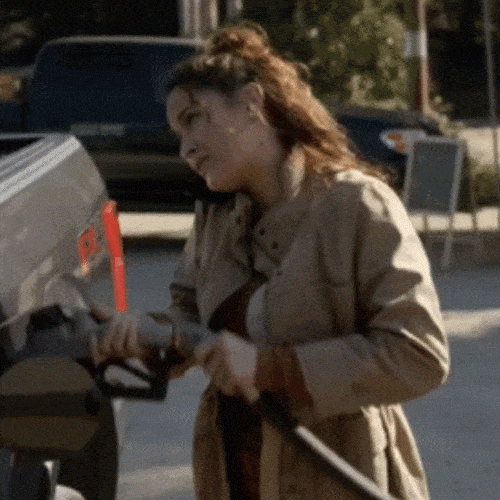 A woman in a coat holds a gas pump nozzle while fueling a vehicle.