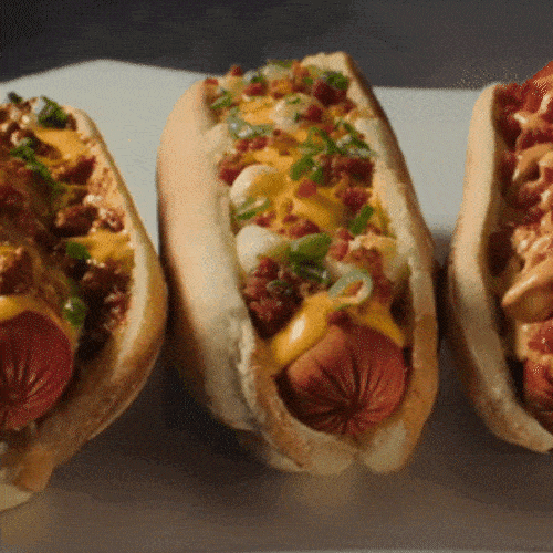 Three hot dogs in buns topped with cheese sauce, chili, and green onions sit side by side.