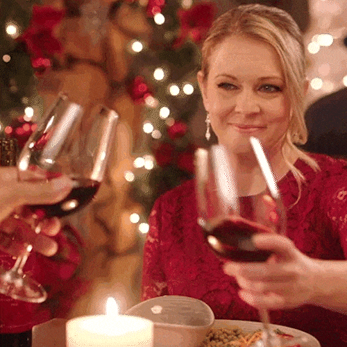 A woman in a red dress raises a glass of red wine at a candlelit dinner by a fireplace.